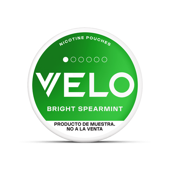 Un envase redondo VELO Mini Trial con una etiqueta VELO verde que dice Bright Spearmint y "bolsa de nicotina sin tabaco". Debajo, "PRODUCTO DE MUESTRA. NO A LA VENTA" indica que es un producto de muestra no a la venta.