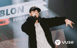 Un artista con chaqueta y gorra negras rapea en el escenario con gestos dinámicos de las manos. Las luces brillantes del escenario y un letrero borroso en el fondo dicen "España". El logotipo de Vuse aparece en la esquina inferior derecha.