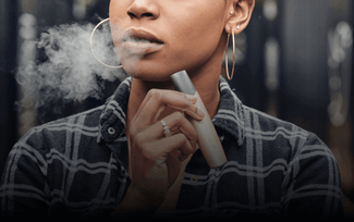 Una persona con pendientes de aro y camisa de cuadros exhala vapor mientras sostiene un dispositivo de vapeo cerca de la boca.